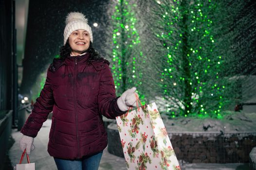 Delightful Latin American woman walking with shopping bags on a city street, illuminated by garlands at snowy winter night. Christmas and New Year concept