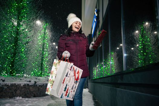 Cheerful multi-ethnic woman with a present box and shopping bags, walking down the city street illuminated by garlands, in the snowy winter evening. Christmas concept. New Year. Winter holidays