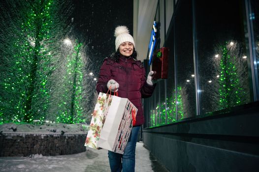 Charming woman with shopping bags and Christmas gifts, walking down the street, lightened by festive illumination at winter snowy night. Christmas holidays. Enjoying winter