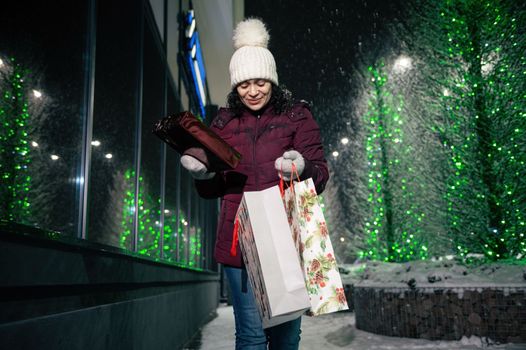 Beautiful woman with shopping bags and Christmas gifts, walking down the street, lightened by festive illumination at winter snowy night. Christmas holidays. Enjoying winter