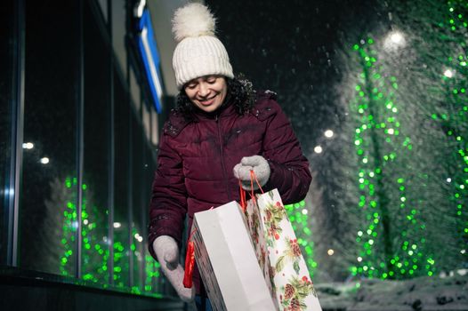 Charming multi-ethnic woman with shopping bags and Christmas gifts, walking down the street, lightened by festive illumination at winter snowy night. Christmas holidays. Enjoying winter