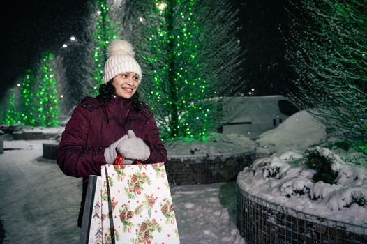 Charming multi-ethnic woman with shopping bags, walking down the city street, lightened by festive illumination at winter snowy night. Christmas holidays. Enjoying winter. Fir-trees with garlands