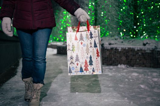 Selective focus on shopping bags with Christmas pattern in the hands of a pretty woman, walking down the snow covered city street, on a winter night, against fir-trees illuminated by festive garlands