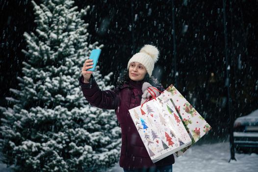 Charming woman taking a selfie on her smartphone while standing with shopping bags on the background of a snow-covered fir tree on a snowy winter evening. Merry Christmas concept