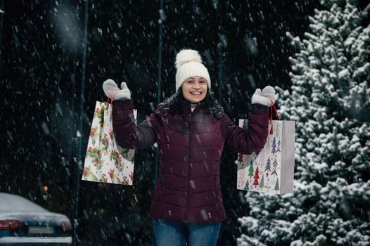 Beautiful multi-ethnic woman in warm clothes, wool hat and mittens, with shopping bags in her hands, smiling a toothy smile, looking at the camera, on a snowy winter evening. Enjoying winter