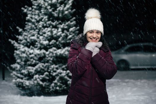 A snow fall on the city street, as a beautiful multi-ethnic woman in white wool hat and mittens, smiling a toothy smile looking at camera, standing against a snow-covered fir-tree on a snowy night