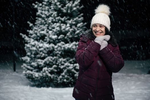 Charming African American woman, in warm clothes, smiling a beautiful toothy smile, having fun standing on a snow covered nature, on a snowy cold winter evening. Christmas and Happy New Year concept