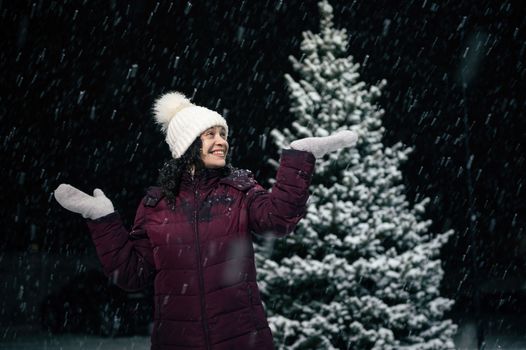 Charming African American woman in white wool hat and mittens, experiencing happiness, catching snowflakes falling on her hands, on a snowy winter evening, standing against a snow covered pine tree