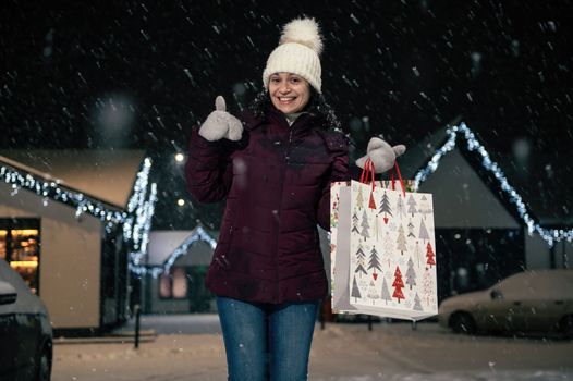 Charming young woman in white wool hat and mittens, experiencing happiness, showing a thumbs up, smiling cheerfully to the camera, standing against a snow covered pine tree on a snowy winter evening