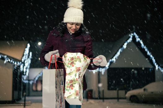 Multi ethnic pretty woman wearing dawn jacket, wool mittens and hat, with shopping bags walking down the snow covered street on a snowy winter cold evening. Merry Christmas and Happy New Year concept