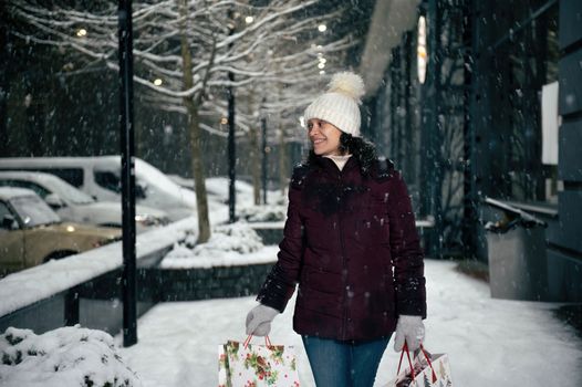 Delightful Hispanic woman in warm clothes, walking down the city street, illuminated by garlands, with shopping bags in her hands, on a snowy winter night. Merry Christmas and Happy New Year concept