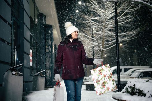 Delightful young multi-ethnic woman walking down the street with shopping bags, on a snowy winter cool evening. Merry Christmas.