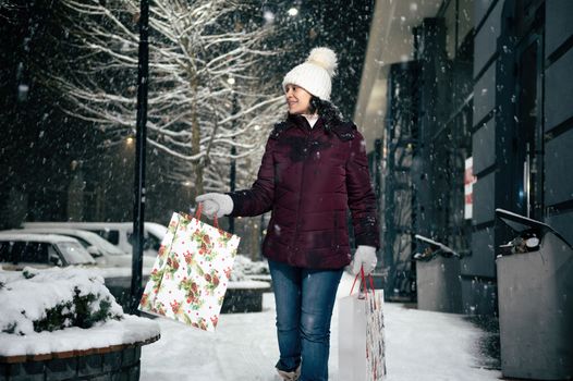 Delightful happy young multi-ethnic woman walking down the street with shopping bags, on a snowy winter cool evening. Merry Christmas.