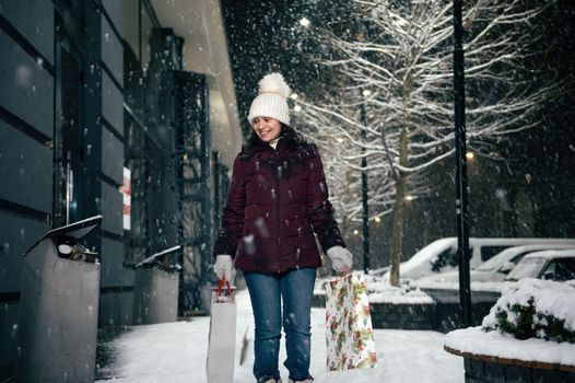 Delightful young multi-ethnic woman walking down the snow covered city street, with shopping bags with Christmas gift box in her hands. Merry Christmas. Snowfall. Winter holidays concept
