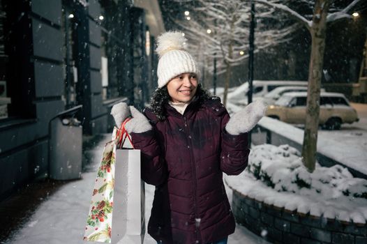 Attractive multi-ethnic young woman in warm clothes, with shopping bags, catches snowflakes falling on her hands while walking along a snowy street illuminated by holiday garlands. Merry Christmas
