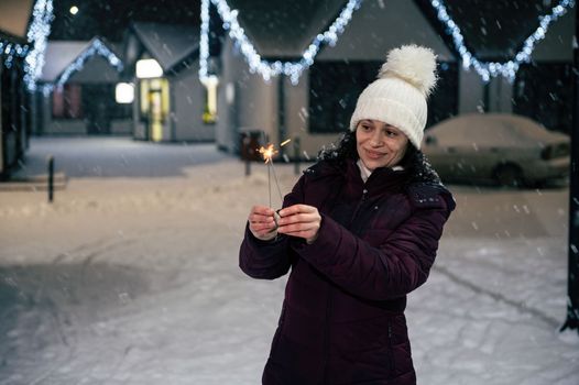 Pretty multi-ethnic woman lighting sparklers, Bengal lights while walking on a city street, illuminated by garlands on a snowy evening. Enjoying winter and snow covered urban environment