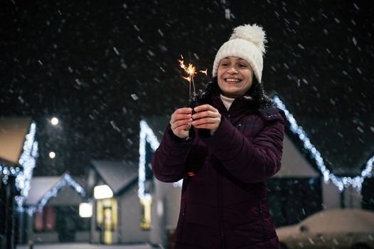 Delightful woman lighting sparklers, Bengal lights on a city street, illuminated by garlands on a snowy evening. Merry Christmas concept. Winter holidays.