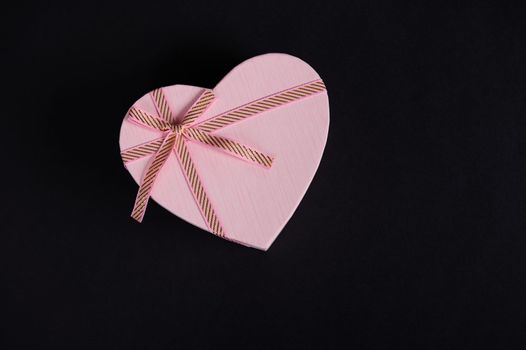 Still life. Flat lay composition with a heart shaped pink gift box with tied bow, isolated on black background with copy advertising space. Romantic present for Saint Valentine's or Women's Day