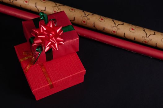 Still life with gift boxes, ornate with green ribbon and red tied bow, surrounded by wrapping paper on a black background. Copy space. Happy New Year. Merry Christmas. Winter holidays. Christmastime