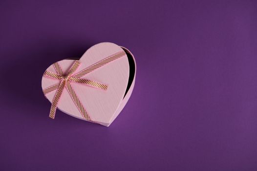 Still life. Top view composition with a half open heart shaped pink gift box with tied bow, isolated on violet background with copy ad space. Romantic present for Saint Valentine's or Women's Day