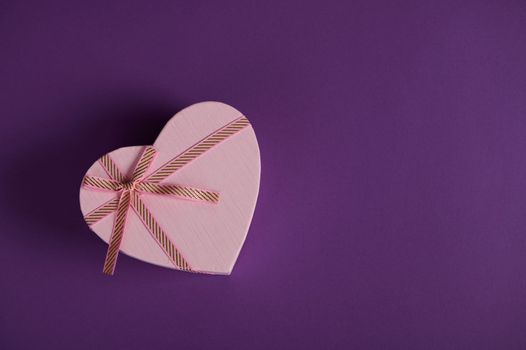 Flat lat with a Saint Valentine's day or International Women's Day heart shaped gift box, with pink ribbon bow, on purple background with copy advertising space. March 8. February 14. Anniversary gift