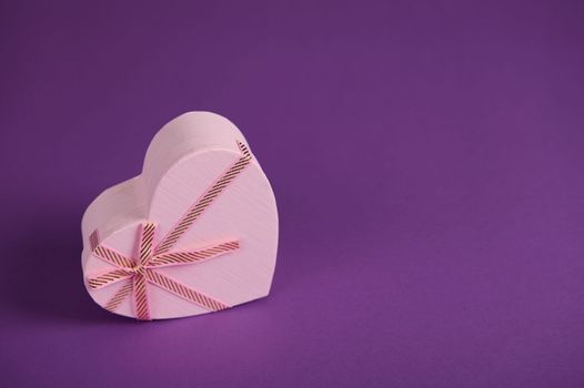 Still life with a pink heart shaped gift box, with a tied bow and romantic surprise inside, isolated in bright purple background. St Valentine's Day. International Women's and Mother's Day. Boxing Day