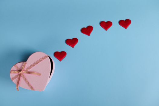 Flat lay. Still life with a pink heart shaped gift box and red decorative hearts, isolated on pastel blue background with copy advertising space. Romantic surprise for St Valentine's Day, Women's Day