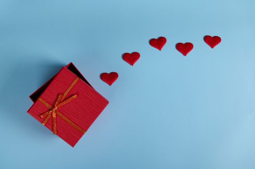 Flat lay. Still life Romantic surprise inside a half open red gift box with tied golden bow and red decorative hearts, isolated on pastel blue backdrop with copy advertising space. St Valentine's Day