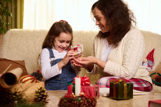 Adorable little child girl enjoys happy winter holidays, helps her loving mother packing beautiful presents for Christmas at cozy home interior. New Year's preparations. Magic festive atmosphere