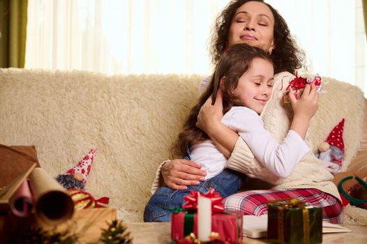 Charming little girl, beloved daughter gently leans on the shoulders of her mother hugging her, receiving a cute Christmas present. Beautiful gifts on the table in foreground. Happy carefree childhood