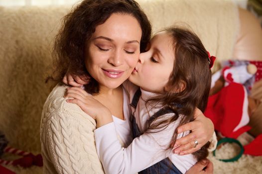 Adorable European little child, cute baby girl, lovely daughter hugging and kissing her loving mother, in Christmas home interior. Happy family relationships. Moments of tenderness, love and care