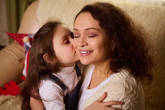 Multi-ethnic beautiful delightful young woman, a loving mother, experiences happiness when her adorable daughter hugs and kisses her on cheek. Happy family relationships. Mother's Day. Children's Day