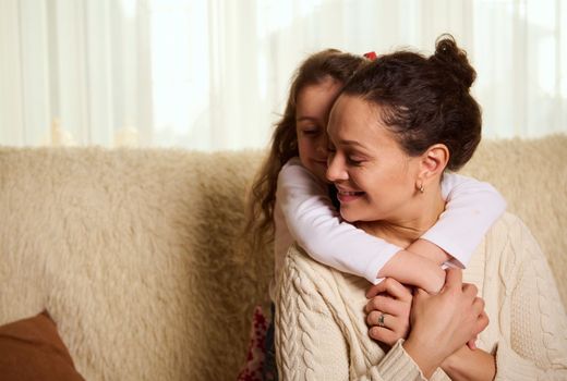Selective focus on a multi-ethnic pretty woman embracing her cute baby girl Beautiful little child, adorable daughter hugging her mum from the back, sitting on sofa at home, showing her love. Family