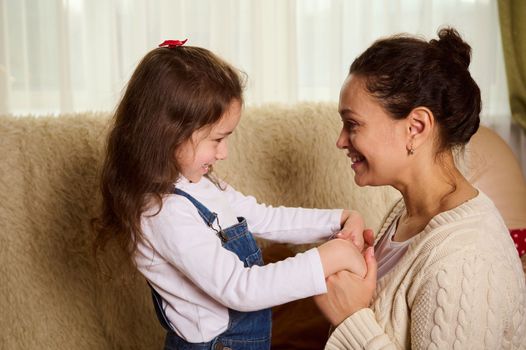 Happy middle-aged pretty woman, a loving mother and her little child daughter smiling, hugging, having fun, spending leisure time together, sitting on couch at home. Two generations. Good relationship