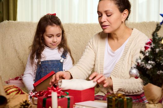 Happy mother and daughter packing presents for Christmas or New Year, looking forward at the upcoming winter holidays. Happy family relationships. Magical festive atmosphere at cozy home interior