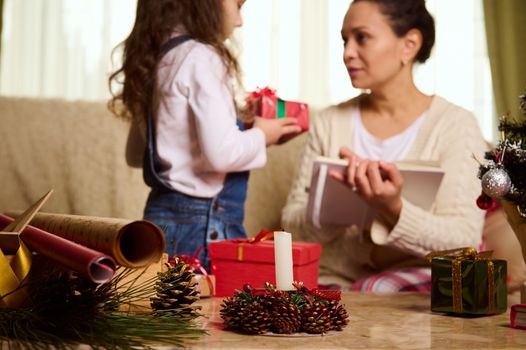 Focus on a candle, wrapping materials and pine cones on blurred background of beautiful child, adorable baby girl in blue denim overalls gives a cute Christmas present to her loving mother. New Year