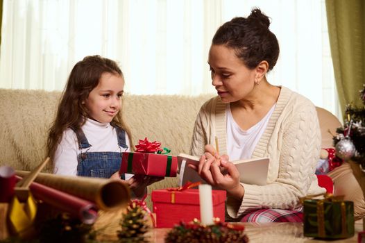 Beautiful multi-ethnic woman, a happy loving mother and her lovely little child girl, adorable daughter, make together a list of gifts, wrapping presents for Christmas or New Years. Winter holidays