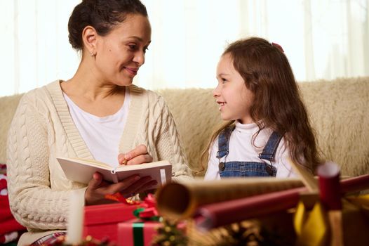 Happy family concept. Loving affectionate mother and her lovely charming daughter make Christmas gift list, smile happily, looking at each other, wrap presents, spend time together on winter holidays