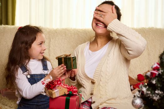 Beautiful woman, a loving mother smiling a cheerful toothy smile, closing her eyes with her hand while her adorable daughter gives her a cute Christmas present. Magical atmosphere of winter holidays