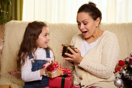 Delightful loving mother rejoices while receive a Christmas present from her adorable little daughter. Lovely baby in blue denim overalls gives a cute Xmas gift box to her caring mom. Happy New Year