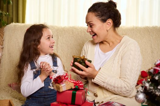 Beautiful woman, a loving mother and her adorable child, a cute little daughter express positive emotions, rejoicing while exchanging Christmas gifts, enjoying happy winter holidays together at home