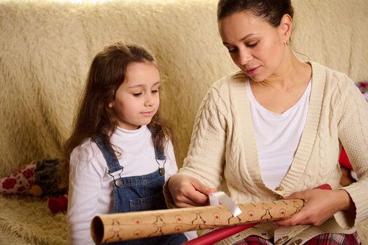 A happy mother and her daughter choose a decorative ribbon, put it over a deer pattern wrapping paper, packing Christmas New Year presents and gift boxes. In expectancy for winter events