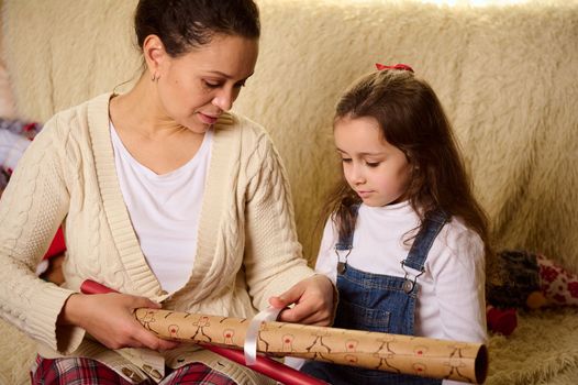 Young mother and little girl daughter using paper for wrapping packing Christmas New Year presents, preparing gift boxes for celebration holiday event at home together.