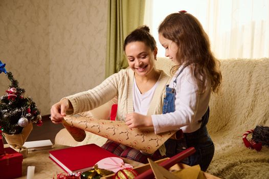 Pretty woman, loving affectionate mother and little girl daughter using wrapping paper for packing Christmas New Year presents, preparing gift boxes for celebration holiday event at home together.