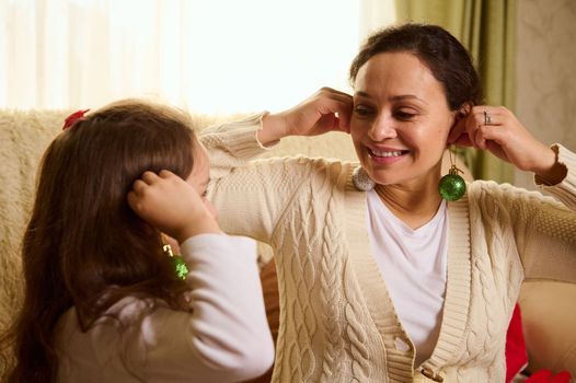 Beautiful multi-ethnic woman wearing warm beige sweater, holding shiny balls as earrings near her ears, have fun while decorate Christmas tree with her daughter, enjoying New Year's preparations
