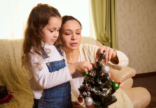 Affectionate loving mother and lovely daughter decorating a Christmas tree together, looking forward to the upcoming winter holidays. Happy winter holidays in family at home. New Year's celebration
