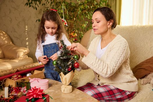Happy multi-ethnic family - Latin American pretty woman, a loving caring affectionate mother and her lovely daughter , a little child girl decorating Christmas tree at home. Gift boxes on foreground