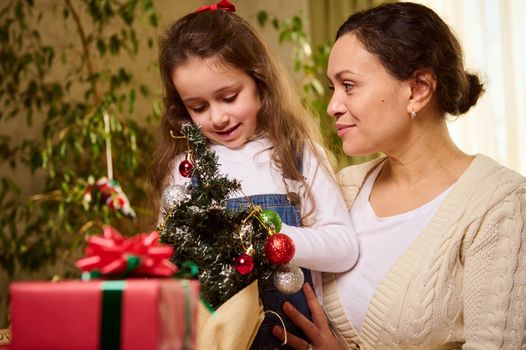 Beautiful young woman, a loving affectionate mother hugging her adorable little daughter, who is hanging toy balls and bauble on a small Christmas tree, making preparations for celebrating New Year