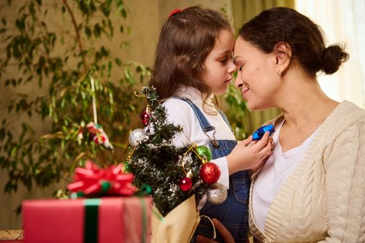 Happy woman, loving affectionate mother and her beloved little daughter enjoying togetherness, hugging each other while decorating Christmas tree at home. Blurred beautiful presents on the foreground