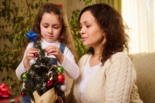 Adorable Caucasian little girl hangs toys on Christmas tree, standing next to her loving mother, in cozy living room. Gorgeous woman decorates home with her lovely daughter for upcoming winter events
